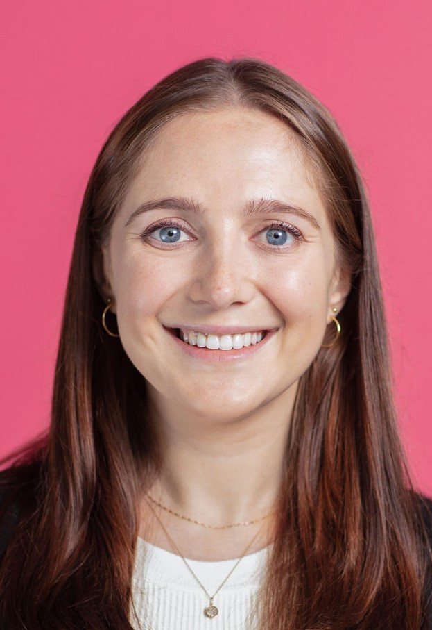 A woman with long brown hair smiling in front of a pink background.